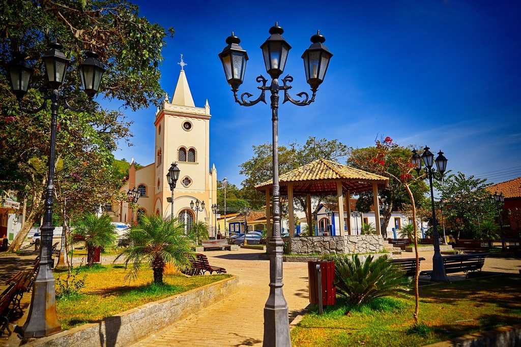 Praça com postes de ferro, coreto e jardins diante da Igreja Matriz de Nossa Senhora da Imaculada Conceição, sob céu azul