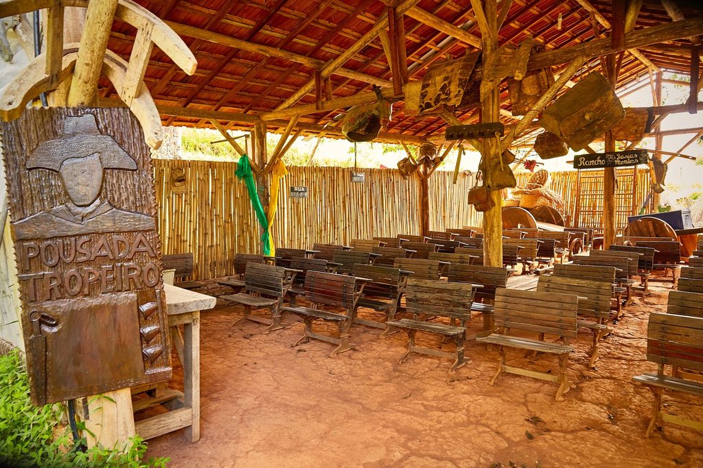 Rancho rústico de madeira com bancos, objetos antigos de tropeiros pendurados e placa entalhada com a figura de um tropeiro