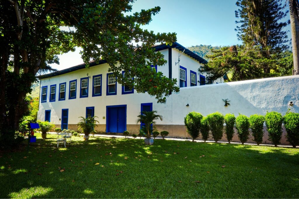 Casarão branco de dois andares da Fazenda São Francisco, com janelas e portas azuis, gramado e árvores, aos pés da serra