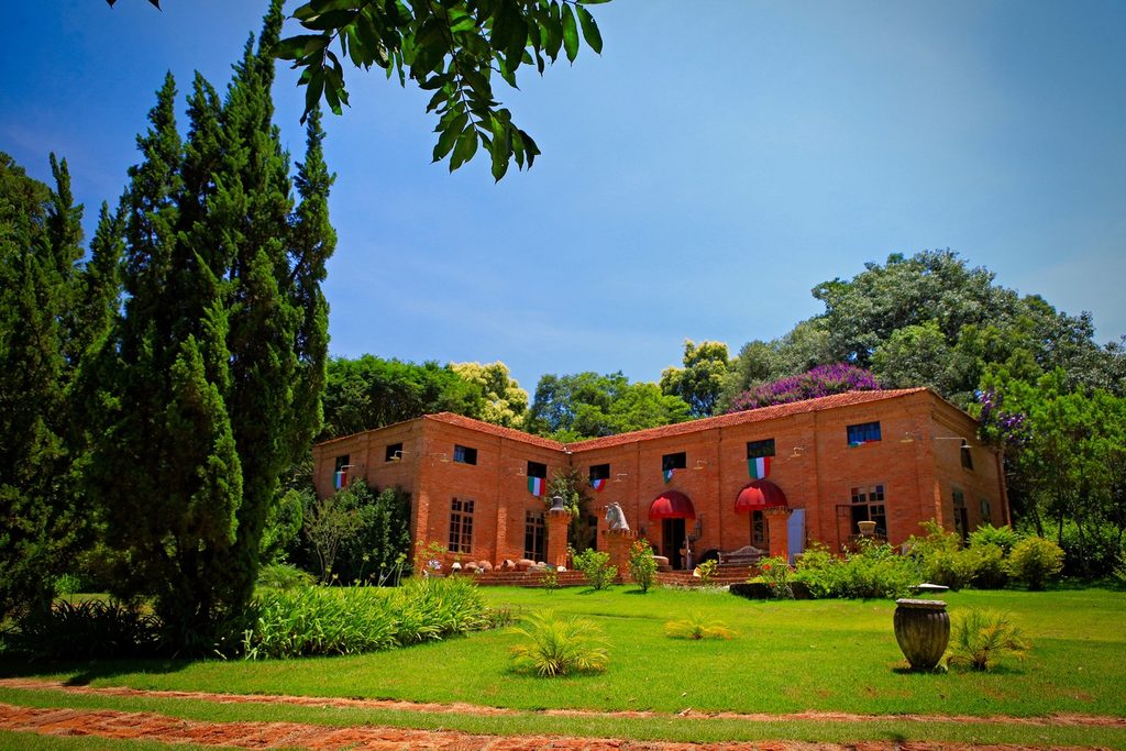 Casarão de tijolos com toldos vermelhos, jardim gramado e ciprestes, sede da Vila del Capo