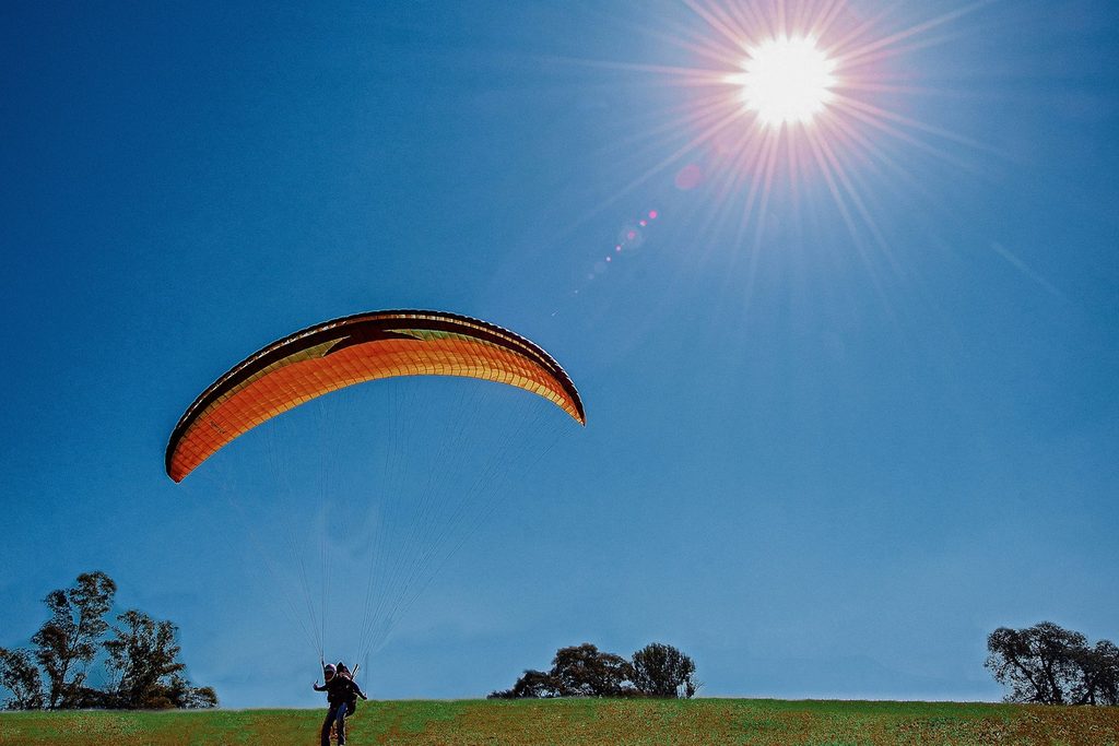 Parapente laranja e preto subindo no céu azul com o sol forte, logo após decolar de um gramado no alto da serra