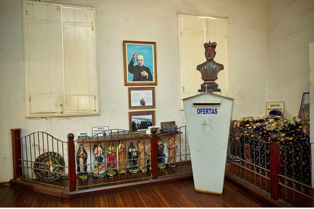 Sala da Casa dos Milagres com busto e retrato do Padre Donizetti, imagens de santos e objetos deixados por fiéis