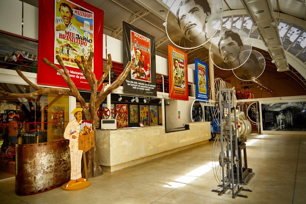 Salão do Museu Mazzaropi, em Taubaté, com cartazes coloridos dos filmes, boneco do Jeca, projetor antigo e retratos do artista pendurados no teto
