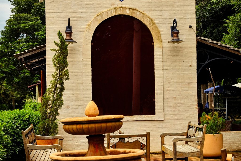 Fachada de tijolos claros com porta em arco, fonte de pedra, bancos de madeira e jardim no distrito de Quiririm