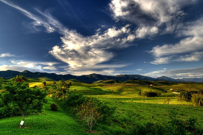 Vista do campo em Taubaté, com morros verdes, árvores e a serra ao fundo sob céu azul com nuvens
