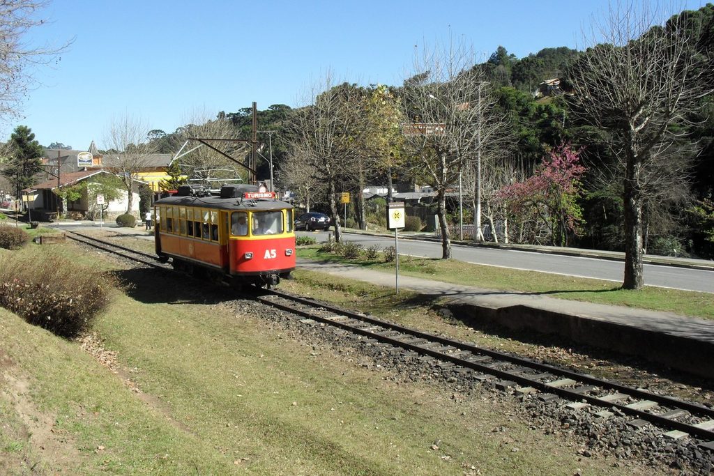 Bondinho A5 do trem de Campos do Jordão passando por trilhos entre gramado, árvores e casas da cidade