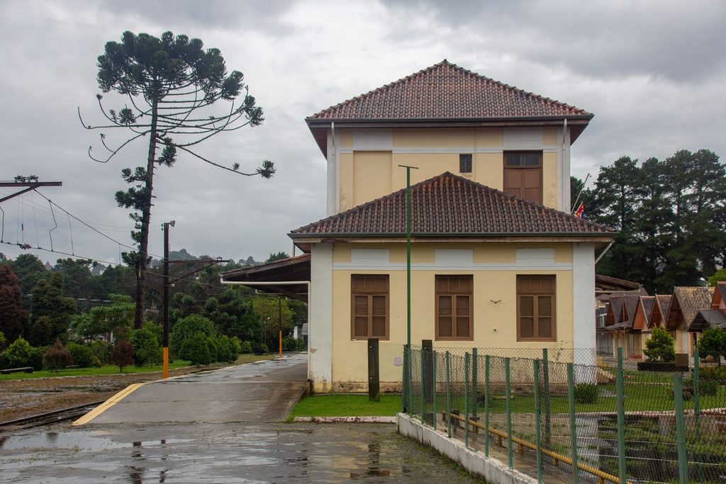 Prédio de estação ferroviária de dois andares, amarelo com telhado vermelho, ao lado de uma araucária