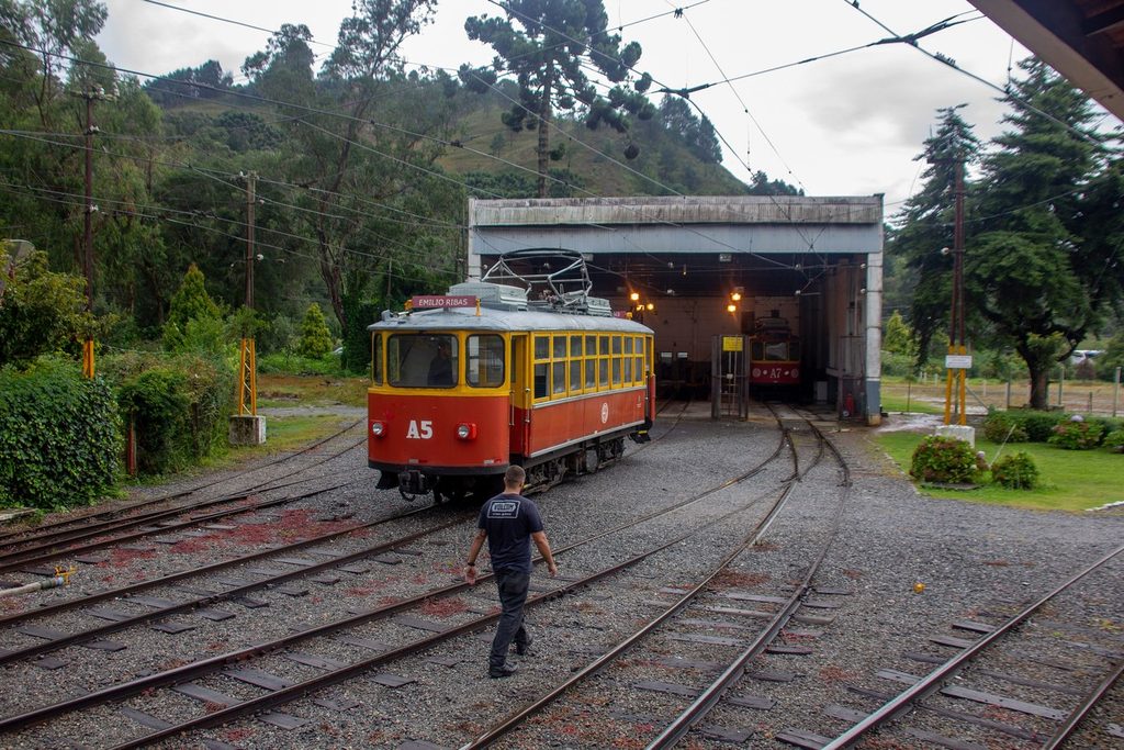 Bondinho A5 do trem de Campos do Jordão saindo da garagem da ferrovia, com trilhos em curva e montanha ao fundo
