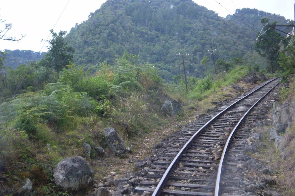 Trilho da Estrada de Ferro Campos do Jordão subindo em curva pela encosta da serra, entre pedras e mato