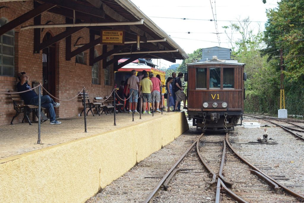 Carro de madeira V1 parado na plataforma coberta de uma estação, com passageiros esperando