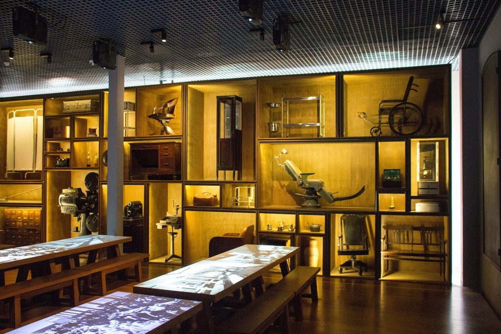 Sala de exposição com móveis, objetos antigos iluminados em nichos de madeira e mesas interativas
