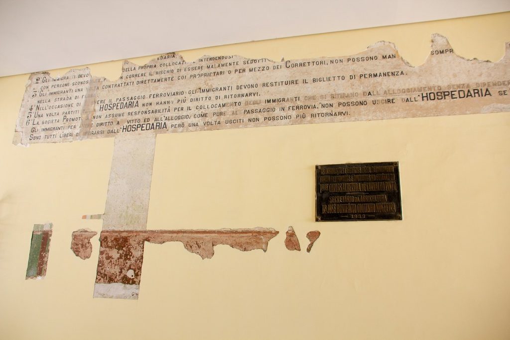 Parede antiga com avisos em italiano pintados, parcialmente descascados, citando a Hospedaria