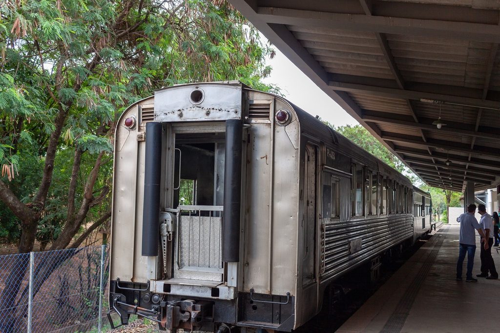 Vagão de passageiros prateado com porta traseira aberta, parado na plataforma coberta da estação, com passageiros conversando