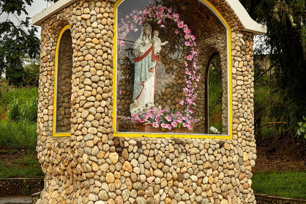 Gruta de pedras redondas com a imagem de Nossa Senhora da Glória enfeitada com flores cor-de-rosa, em Tremembé