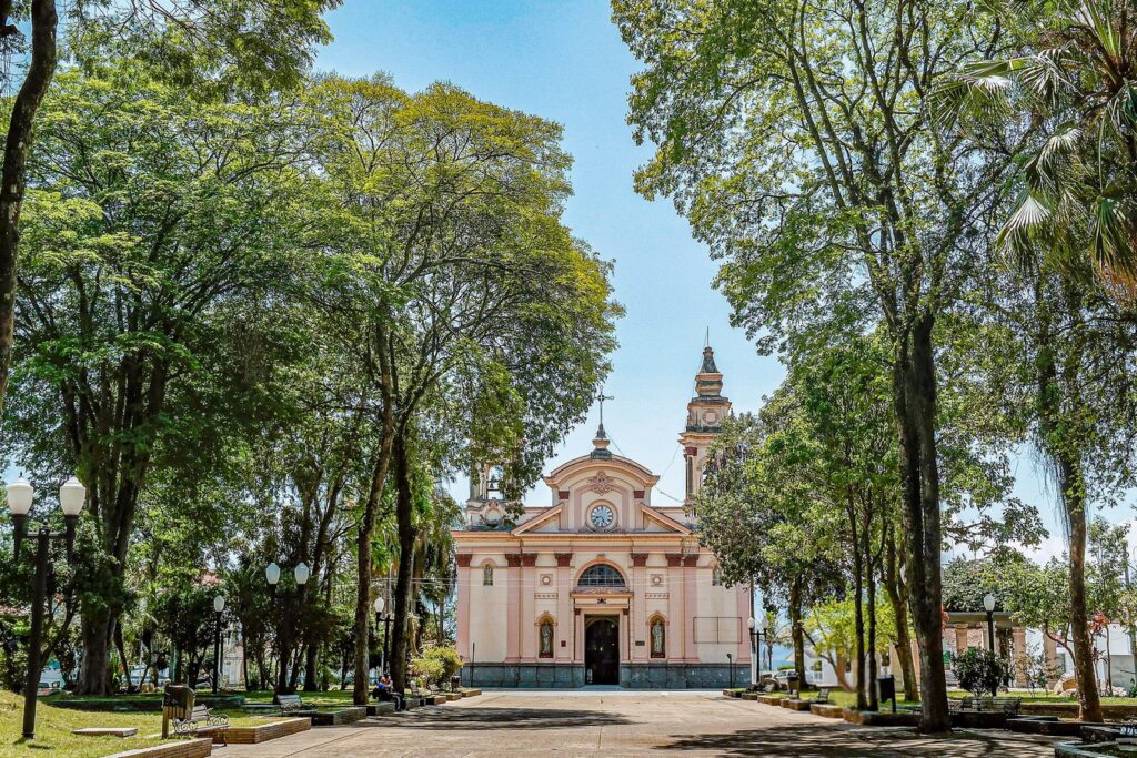 Alameda arborizada da Praça Padre Luiz Balnes levando à fachada rosa e branca da Basílica do Senhor Bom Jesus de Tremembé