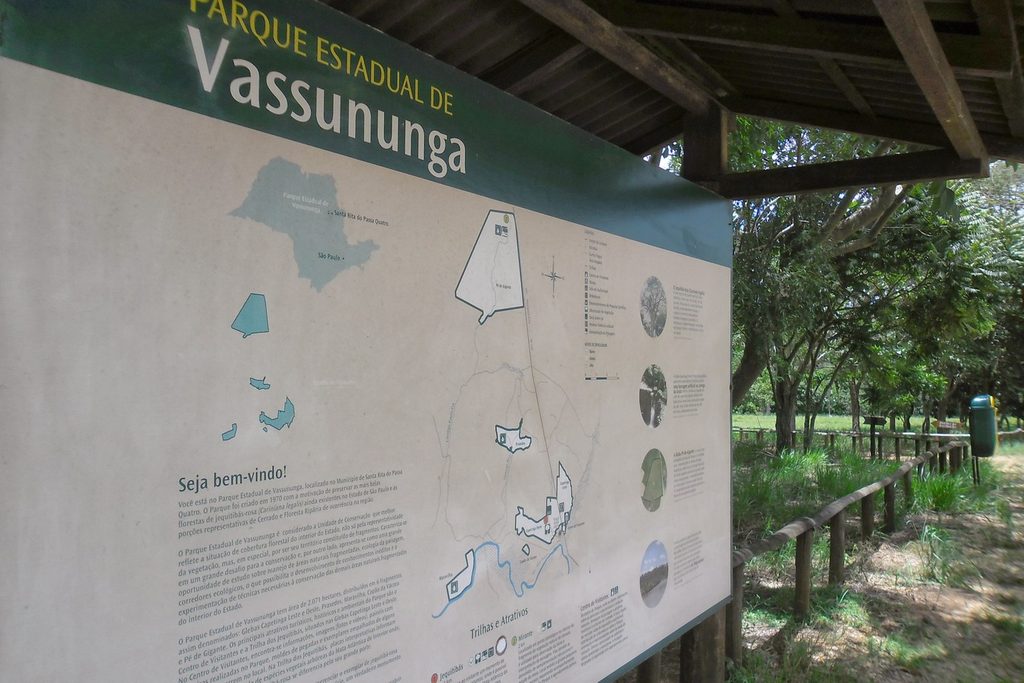 Placa de boas-vindas do Parque Estadual de Vassununga com o mapa das áreas do parque, ao lado de uma trilha com cerca de madeira