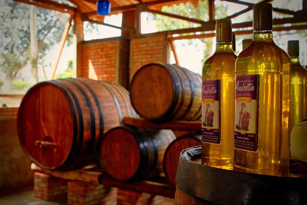 Barris de madeira e garrafas de cachaça dourada com rótulo do Alambique Andretta, num galpão rústico de tijolos
