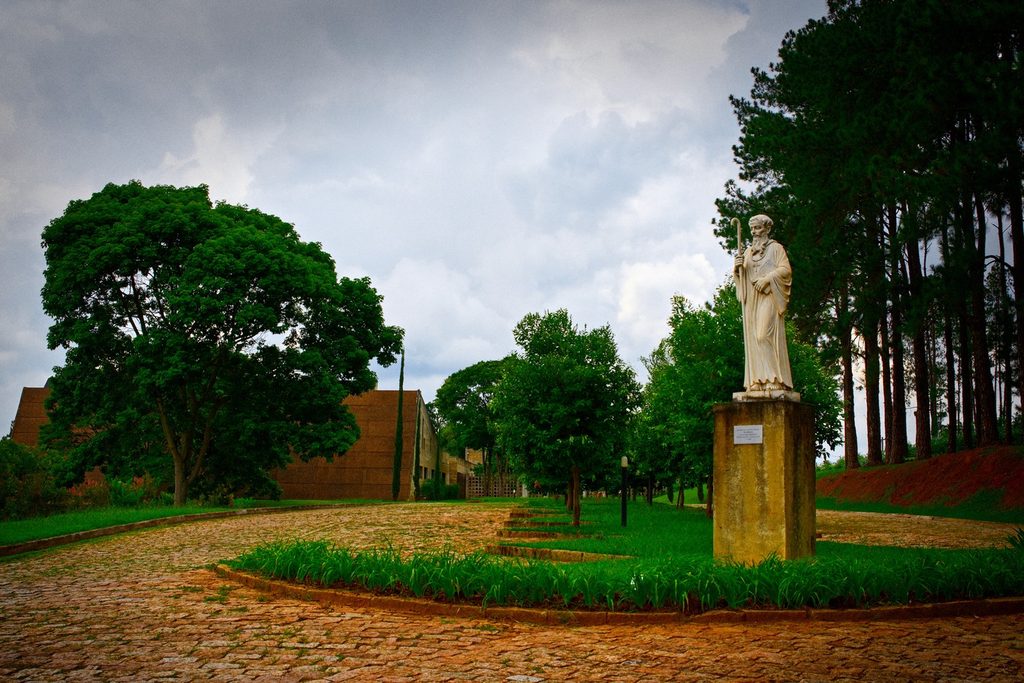 Estátua branca de São Bento num jardim com caminho de pedras e árvores altas, na entrada do Mosteiro de São Bento